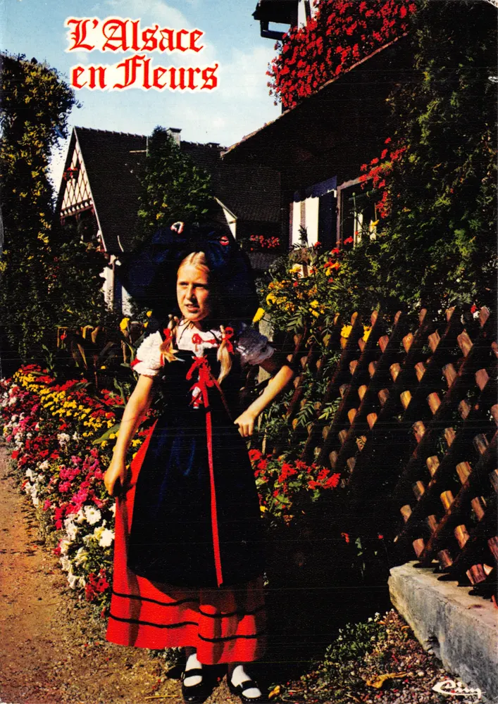 Mädchen im elsässischen Kostüm — vor blühendem Garten und Holzzaun — Editions Hansi - Vorderseite