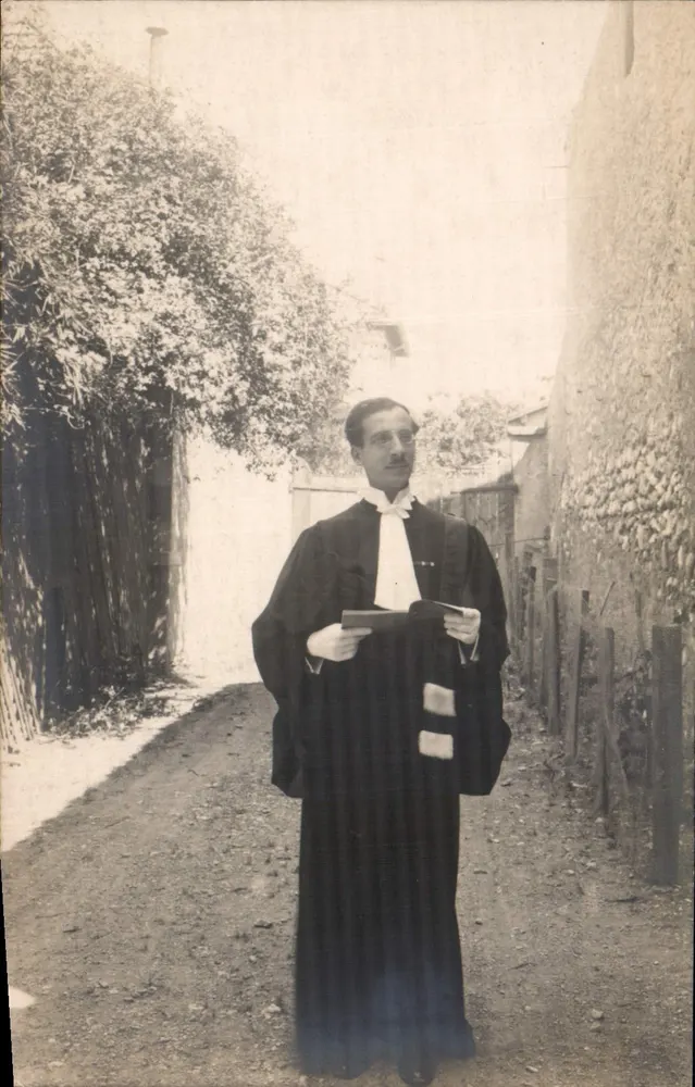Un homme en robe de magistrat — rue pavée entre mur et haie — sans éditeur ni numéro visible - Recto