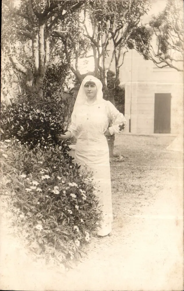 Schwester — in weißem Uniform mit rotem Kreuz — vor einem Garten und einem Gebäude - Vorderseite