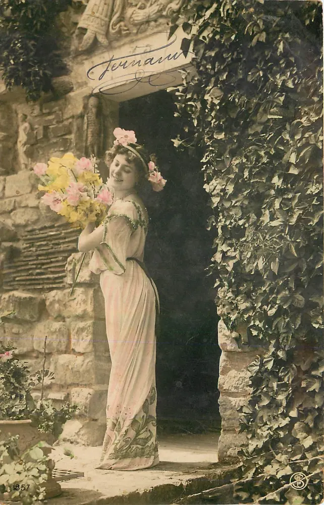 Mulher em vestido de estilo Art Nouveau segurando um buquê — diante de uma porta de pedra e trepadeiras — editada por Jernam, n° 1857 - Frente