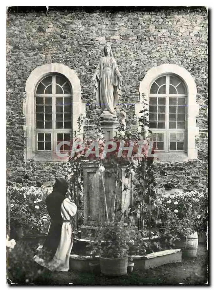Notre-Dame des Neiges (Ardèche) — Die Jungfrau des Cloisters, Statue vor einer Steinmauer mit zwei Bogenfenstern, eine Nonne in schwarz-weißem Kleid knieend vor einem Blumenbecken - Vorderseite