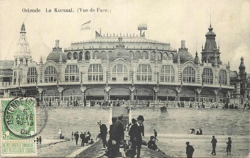 Ostende — O Kursaal. (Vista da Frente.) — Vista do edifício do Kursaal em Ostende, com bandeiras e passeio na praia. - Frente