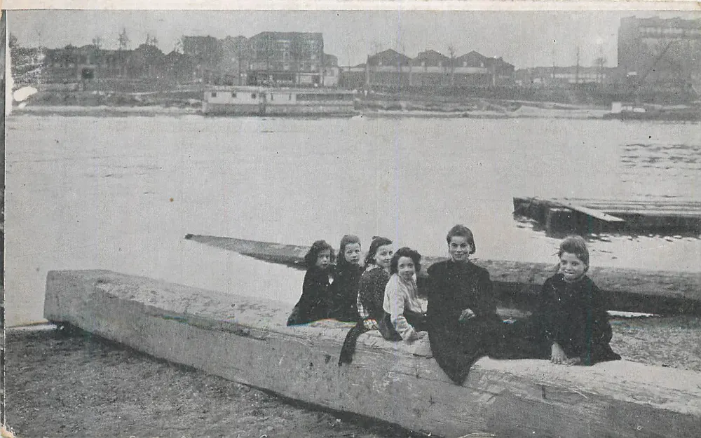Cinco crianças sentadas em um tronco de árvore — margem fluvial com edifícios no fundo — editor não visível - Frente