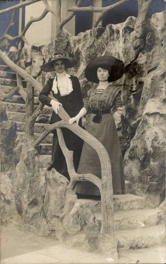Duas mulheres em traje da época — diante de um cenário de rochedos e galhos — cartão postal - Frente