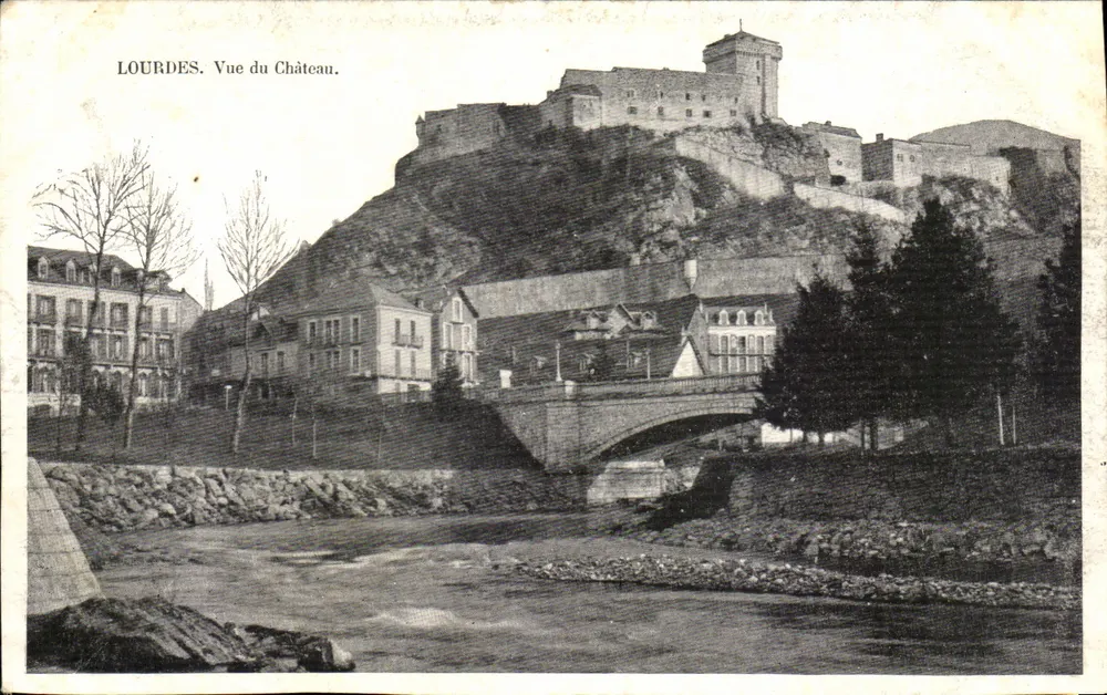Lourdes. Vista do Castelo — vista panorâmica do castelo sobre uma colina, com um ponte e um curso d'água embaixo. - Frente