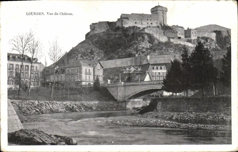 Lourdes. Vista do Castelo — vista panorâmica do castelo sobre uma colina, com um ponte de pedra no primeiro plano e edifícios em primeiro plano. - Frente