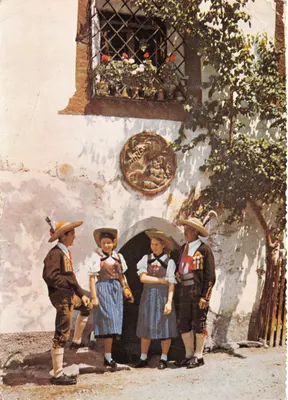 Ötztaler Tracht — scène de rue avec quatre personnes en costume traditionnel devant un mur blanc orné d'un blason et d'un balcon fleuri — Tyrolia-Druck 9471, Nr. 27