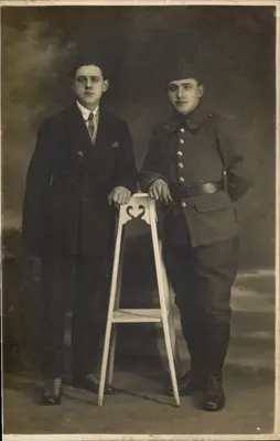 Dois jovens homens — um em traje civil, outro em uniforme militar — posando diante de um banco, estúdio — Carta postal